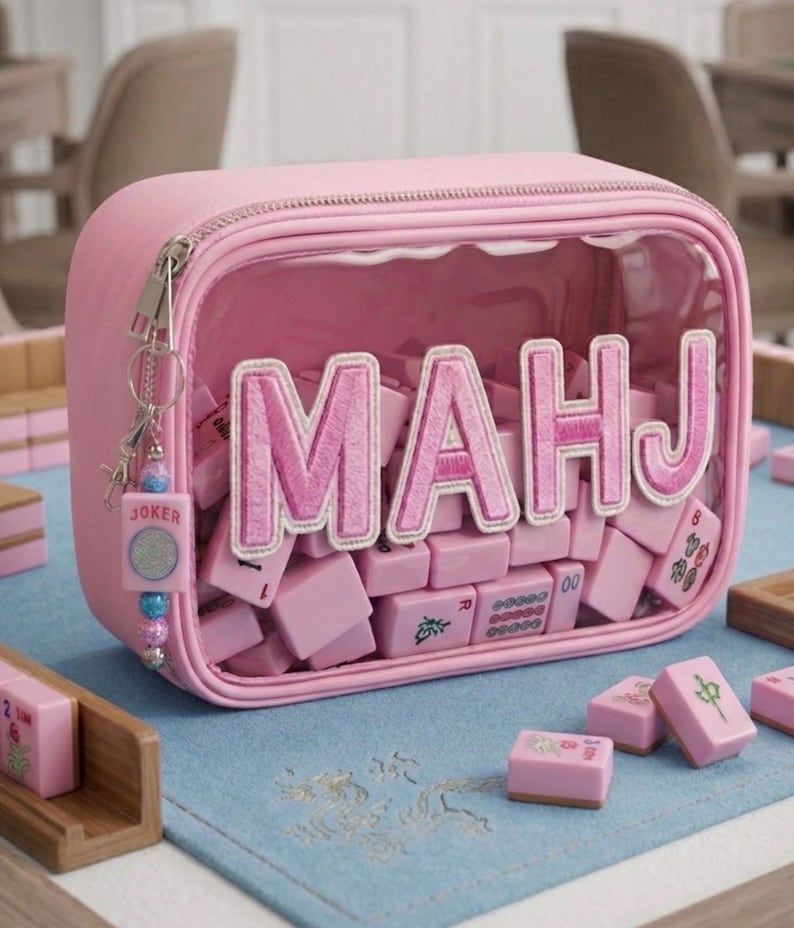 May include: A pink zippered case with a clear front panel, filled with pink Mahjong tiles. The word "MAHJ" is embroidered in pink on the clear panel. A "JOKER" charm hangs from the zipper. The tiles are arranged on a light blue surface.