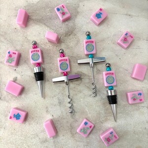 May include: Pink bottle stoppers and corkscrews with a Mahjong theme. The stoppers and corkscrews have a silver finish and are decorated with pink tiles that say "Joker". Pink Mahjong tiles are scattered around the stoppers.