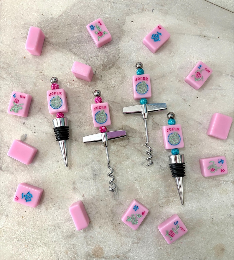 May include: Pink bottle stoppers and corkscrews with a Mahjong theme. The stoppers and corkscrews have a silver finish and are decorated with pink tiles that say "Joker". Pink Mahjong tiles are scattered around the stoppers.