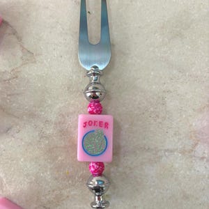 May include: A silver fork with a decorative handle. The handle features a pink square tile with the word "JOKER" and a circular design, along with pink beads and silver accents. The fork is likely for serving appetizers.