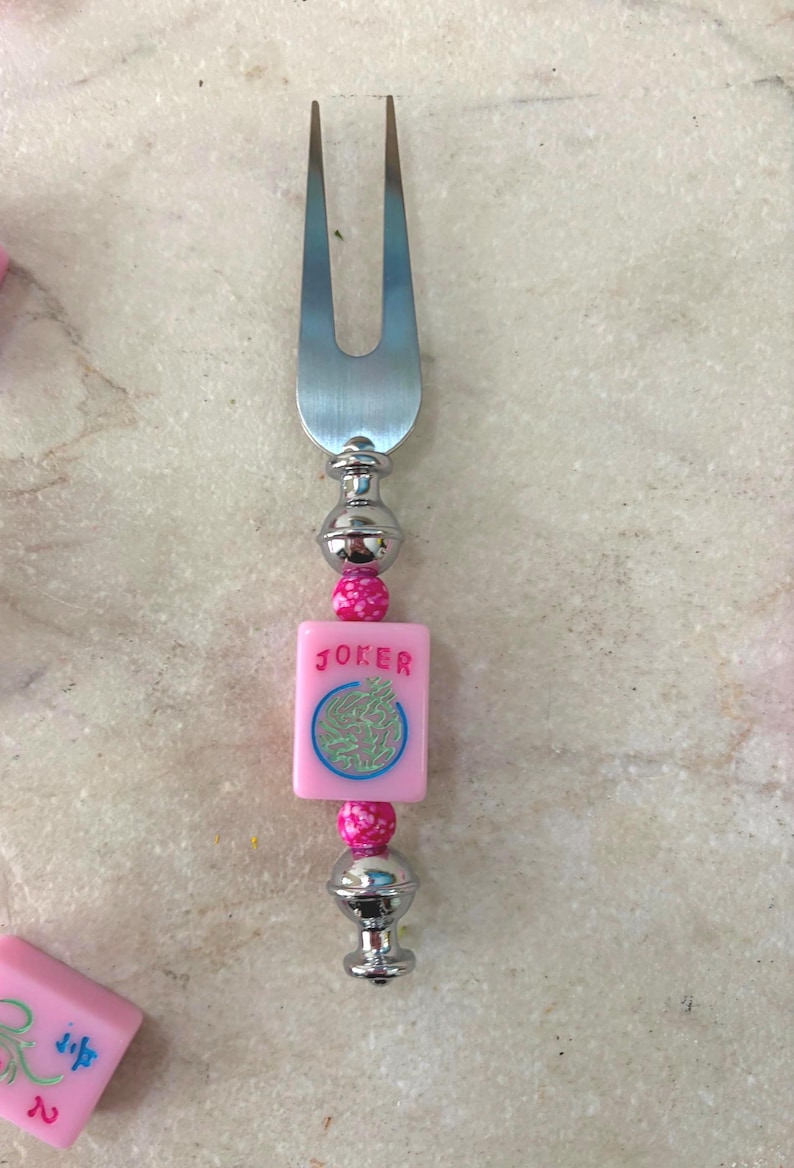 May include: A silver fork with a decorative handle. The handle features a pink square tile with the word "JOKER" and a circular design, along with pink beads and silver accents. The fork is likely for serving appetizers.