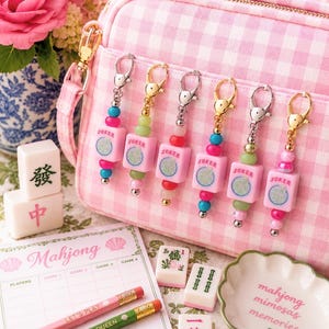 May include: A pink gingham crossbody bag with six matching keychains. Each keychain has a pink square charm with "JOKER" printed on it, and colorful beads. Mahjong tiles, pencils, and a decorative dish are also visible.