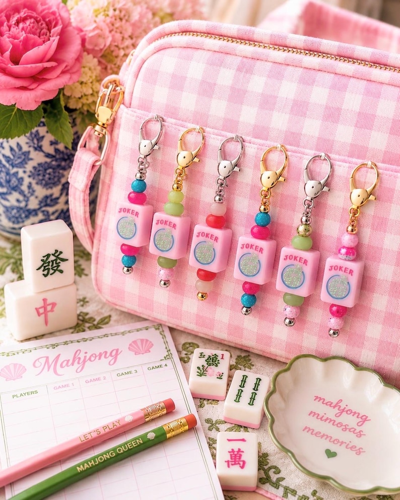 May include: A pink gingham crossbody bag with six matching keychains. Each keychain has a pink square charm with "JOKER" printed on it, and colorful beads. Mahjong tiles, pencils, and a decorative dish are also visible.
