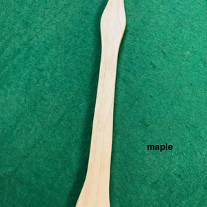 Puede incluir: Una paleta de madera con una punta puntiaguda y un fondo redondeado. La paleta está hecha de madera de arce y tiene una cuerda negra atada a la parte inferior.