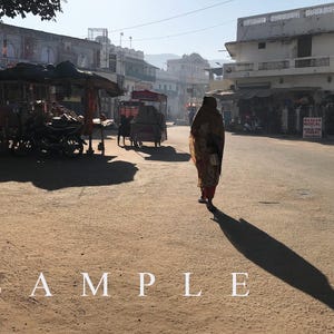 May include: A person wearing a long, patterned garment walks across a sunlit, dusty street in a town with white buildings. The scene features a long shadow cast on the ground, several parked carts, and the word SAMPLE printed in white text.