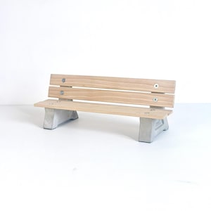 May include: A miniature park bench featuring two light-toned wooden slats for the seat and backrest, secured with four visible silver-colored metal screws. The bench is supported by two solid gray concrete legs with a smooth, matte finish.