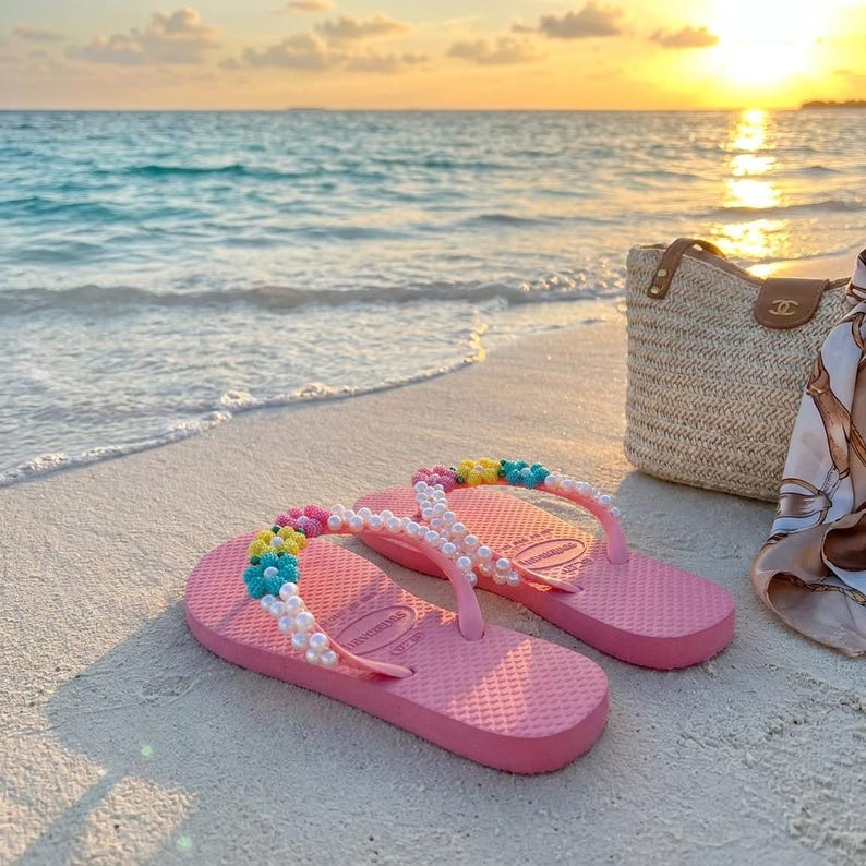 Peut inclure: Une paire de tongs roses avec des brides orn&eacute;es de fausses perles blanches et de petites perles en forme de fleurs color&eacute;es. Les semelles pr&eacute;sentent une texture &agrave; motif quadrill&eacute; classique et le nom de la marque est grav&eacute; au niveau du talon.