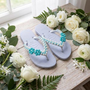 Peut inclure: Une paire de tongs bleu clair orn&eacute;es de perles blanches et de fleurs en perles turquoise sur les brides. Les sandales sont pr&eacute;sent&eacute;es avec des roses blanches et du feuillage vert, mettant en valeur une semelle &agrave; la texture granuleuse.