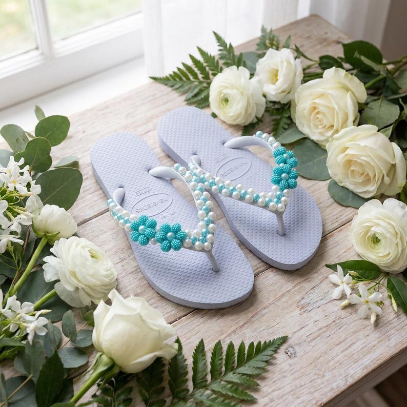 Peut inclure: Une paire de tongs bleu clair orn&eacute;es de perles blanches et de fleurs en perles turquoise sur les brides. Les sandales sont pr&eacute;sent&eacute;es avec des roses blanches et du feuillage vert, mettant en valeur une semelle &agrave; la texture granuleuse.
