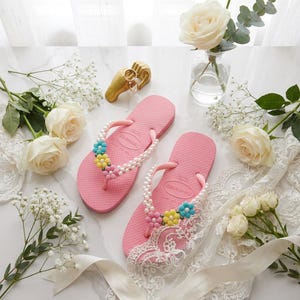 Peut inclure: Une paire de tongs roses orn&eacute;es de perles blanches et de petites d&eacute;corations en forme de fleurs bleues, jaunes et roses sur les brides. L'une des tongs est partiellement recouverte d'un morceau de tissu en dentelle blanche.