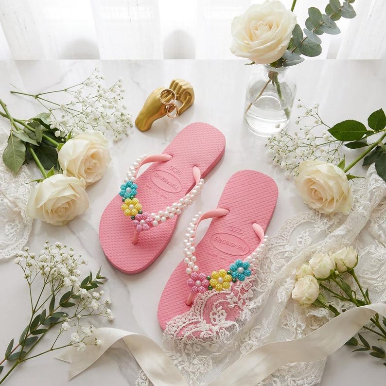 Peut inclure: Une paire de tongs roses orn&eacute;es de perles blanches et de petites d&eacute;corations en forme de fleurs bleues, jaunes et roses sur les brides. L'une des tongs est partiellement recouverte d'un morceau de tissu en dentelle blanche.