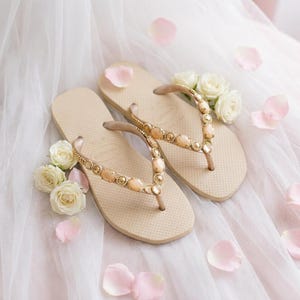 Peut inclure: Une paire de tongs beiges dot&eacute;es de brides orn&eacute;es de cabochons ovales couleur p&ecirc;che clair et de petits accents m&eacute;talliques dor&eacute;s. La semelle pr&eacute;sente une surface textur&eacute;e et le nom de la marque est grav&eacute; au niveau du talon.