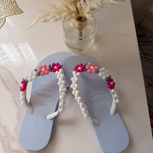 Peut inclure: Une paire de tongs Havaianas bleu clair avec des brides en caoutchouc blanc orn&eacute;es de perles d&eacute;coratives en forme de fleurs roses, blanches et vertes, ainsi que des rang&eacute;es de fausses perles blanches fix&eacute;es sur les brides.