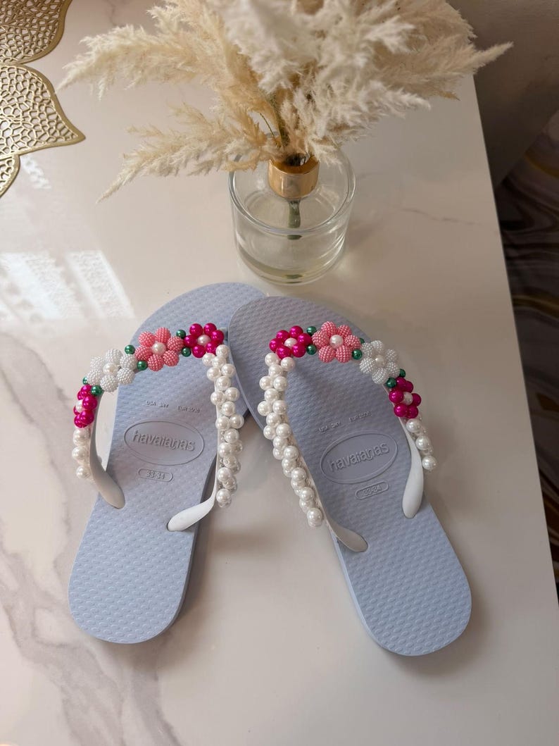 Peut inclure: Une paire de tongs Havaianas bleu clair avec des brides en caoutchouc blanc orn&eacute;es de perles d&eacute;coratives en forme de fleurs roses, blanches et vertes, ainsi que des rang&eacute;es de fausses perles blanches fix&eacute;es sur les brides.