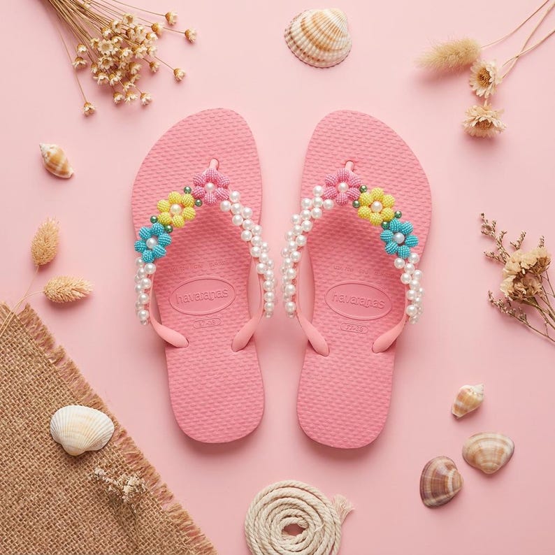 Peut inclure: Une paire de tongs en caoutchouc rose avec la marque Havaianas sur la semelle. Les brides sont orn&eacute;es de fausses perles blanches et de petites fleurs perl&eacute;es jaunes, roses et bleues. Les semelles textur&eacute;es pr&eacute;sentent un design classique.