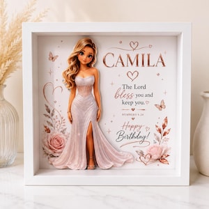 Könnte beinhalten: Ein weißer Schattenfugenrahmen mit einer 3D-Illustration einer blonden Figur in einem schimmernden hellrosa Kleid mit Seitenschlitz. Der Hintergrund zeigt rosa Rosen, Schmetterlinge und den Text CAMILA, The Lord bless you and keep you, Happy Birthday.