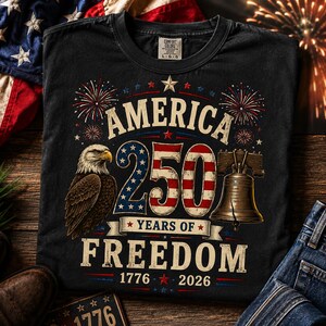 K&ouml;nnte beinhalten: Ein schwarzes T-Shirt mit einer Grafik, die einen Wei&szlig;kopfseeadler, die Liberty Bell und Feuerwerk zeigt. Der Text lautet America 250 Years of Freedom 1776 2026. Die Zahl 250 ist mit Sternen- und Streifenmustern in Rot, Wei&szlig; und Blau gestaltet.