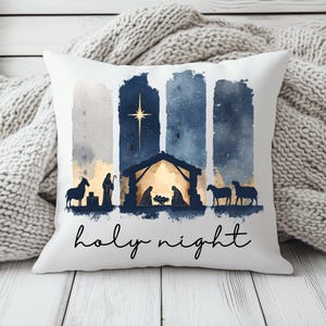 Könnte beinhalten: Ein weißes quadratisches Dekokissen mit einer Krippenszene im Aquarellstil in Marineblau und Gold. Das Motiv zeigt einen Stall, Figuren und Tiere unter einem hellen Stern, mit dem schwarzen, kursiven Schriftzug holy night darunter.