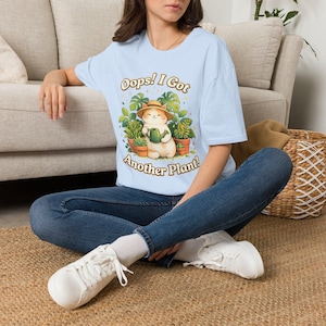 May include: A light blue t-shirt featuring a graphic of a cat wearing a straw hat and holding a green watering can, surrounded by potted plants. The text "Oops! I Got Another Plant!" is printed in brown letters above and below the illustration.