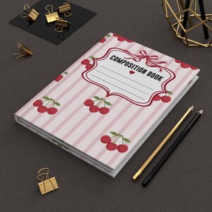 May include: A composition book with a light pink and white vertical striped cover featuring a repeating pattern of red cherries. The center label has a decorative red border, a small red heart, and the text COMPOSITION BOOK in black.