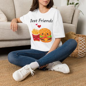 May include: A white short-sleeve t-shirt featuring a graphic of a cartoon hamburger and a carton of french fries with the text "Best Friends" above them, accompanied by two small red hearts, paired with blue denim jeans and white sneakers.