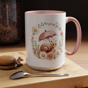 May include: A white ceramic mug with a soft pink interior and handle. The exterior features a graphic of a brown snail holding a pink polka-dot umbrella, surrounded by a circular wreath of white and pink daisies and green foliage.