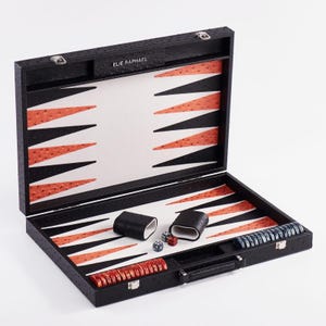 May include: Open black backgammon set with a textured finish, featuring a white and orange triangle design. Includes dice, dice cups, and game pieces in red and blue. The brand name "ELIE RAPHAEL" is printed on the top.
