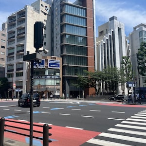 以下が含まれることがあります： ガラス窓と茶色のファサードを備えたモダンな高層ビルがある都市のストリートシーン。赤と白のマーキングのある横断歩道が建物につながっています。黒い車が道路を走っています。