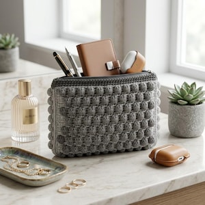May include: A rectangular gray crochet pouch featuring a textured bobble stitch pattern. The pouch is filled with a tan leather notebook, a black pen, a silver pen, and a tan earbud case. The zipper closure is at the top of the pouch.