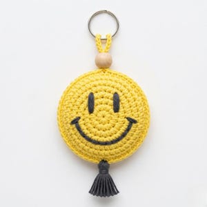 Op de afbeelding: Een ronde, gele gehaakte sleutelhanger met een donkergrijs geborduurd smileygezicht. De sleutelhanger bevat een natuurlijke houten kraal, een zilverkleurige metalen ring aan de bovenkant en een donkergrijs garenkwastje aan de onderkant.