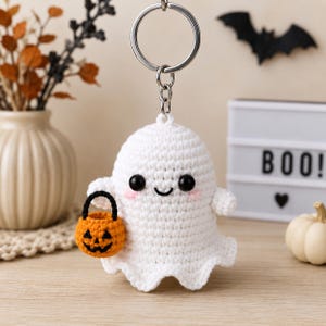 May include: A white crocheted ghost keychain featuring black bead eyes, a stitched smile, and pink cheeks. The ghost holds a small orange crocheted pumpkin bucket with a black handle. The figure is attached to a silver metal keyring and chain.