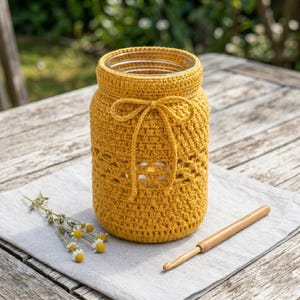Puede incluir: Un portamaceta de ganchillo amarillo mostaza con un lazo decorativo, colocado sobre un paño de lino gris. El portamaceta tiene un diseño calado y va acompañado de un ganchillo de madera. También hay un pequeño ramo de flores blancas y amarillas.