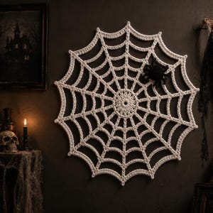 May include: A circular white crocheted spider web wall hanging featuring a small black crocheted spider attached to the upper right section. The web has a central ring with radiating spokes connected by concentric rows of crocheted stitches.