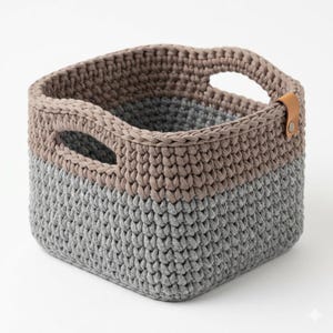 May include: A square crochet basket featuring a two-tone design with a light gray base and a taupe upper section. The basket includes two integrated side handles and a small tan leather-like tab attached to the rim with a silver metal rivet.