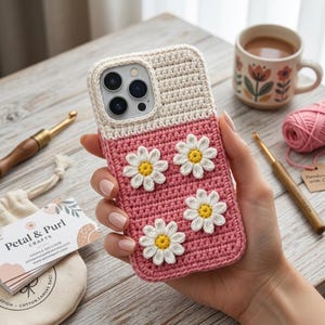 Może przedstawiać: Prostokątne etui na telefon z dwukolorowym wzorem szydełkowym, składające się z kremowej górnej części i różowej dolnej części. Trzy białe aplikacje w kształcie stokrotek z żółtymi środkami są przymocowane do różowego obszaru etui.