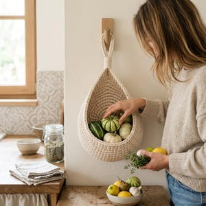 Puede incluir: Una cesta colgante en forma de lágrima de color crema, hecha de cuerda gruesa trenzada. La cesta está suspendida de un gancho de pared de madera y contiene varias calabazas decorativas pequeñas en tonos verdes, blancos y amarillos.