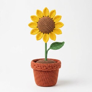 以下が含まれることがあります： 茶色の中心部と鮮やかな黄色の花びらを持つかぎ針編みのヒマワリで、一枚の葉が付いた緑色の茎に繋がっています。花は、質感のあるテラコッタ色のかぎ針編みの鉢に植えられており、縁には装飾が施され、柔らかい布製の植物となっています。