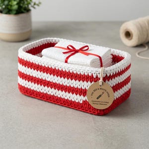 May include: A rectangular crocheted storage basket featuring horizontal red and white stripes. Inside sits a folded white cloth tied with a red ribbon. A round cardboard tag attached to the front reads THE RED & WHITE HOOK, Handmade Crocheted Storage, Washable.