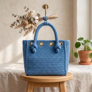 Pode incluir: Uma bolsa tote azul de crochê com ilhós dourados e detalhes de alça com nós. A bolsa é exibida em um banquinho de madeira, com um arranjo de flores secas pendurado acima. A textura da bolsa é visível.