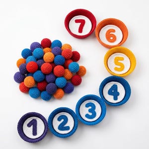 May include: A collection of small, round, crocheted yarn balls in various colors including red, orange, blue, and purple, arranged next to seven circular, crocheted bowls. Each bowl contains a white circular insert with a number from one to seven.