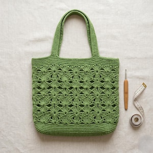 May include: A handmade green crochet tote bag with a floral pattern. The bag has two handles and a rectangular shape. A crochet hook and a measuring tape are next to the bag.
