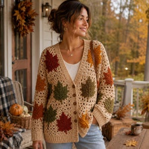 May include: A beige crochet cardigan featuring a V-neckline and a button-down front. The garment is decorated with textured, three-dimensional maple leaf motifs in shades of dark red, burnt orange, and olive green scattered across the fabric.