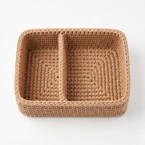 May include: A rectangular, beige crochet basket with a central divider, creating two compartments. The basket is made of thick, textured yarn, with a sturdy, woven appearance. The design is simple and functional, suitable for organizing small items.