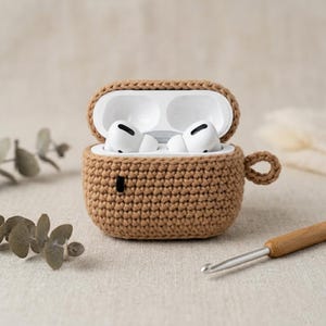May include: A brown, crocheted case for AirPods Pro, with the case open to reveal the white earbuds. The case has a loop for attaching to a keychain. A crochet hook rests nearby.
