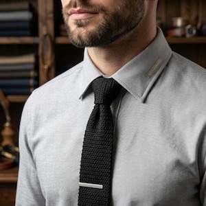 May include: A light gray button-down shirt with a pointed collar featuring a small fabric tag that reads "TEXTILE CRAFTS". The shirt is paired with a black knit necktie and a thin, horizontal silver-toned metal tie bar centered on the tie.