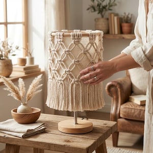 May include: A beige macrame table lamp with a wooden base and a cream-colored shade. The lamp features a woven design and a fringe at the bottom. The lamp is on a wooden table.