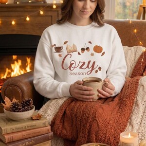 Puede incluir: Una sudadera blanca de cuello redondo con un estampado gráfico que incluye el texto "Cozy Season" en letra cursiva marrón, acompañado de ilustraciones de una taza de café, un libro abierto, bellotas y una calabaza en tonos otoñales.