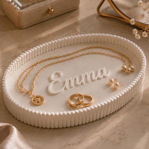 May include: An oval white decorative tray with a ribbed edge featuring the name Emma in raised white script lettering. The tray holds a gold chain necklace with a circular pendant, two gold rings, and a pair of small white pearl stud earrings.