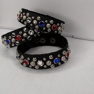 Puede incluir: Tres pulseras de cuero negro con tachuelas plateadas y gemas de colores. Cada pulsera está adornada con gemas rojas, azules y transparentes, creando un contraste llamativo con el cuero negro. Se exhiben sobre una superficie blanca.
