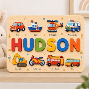May include: A wooden name puzzle featuring the name HUDSON in large, colorful letters. The board includes cutouts for eight vehicles: a car, boat, helicopter, ambulance, excavator, motorcycle, fire truck, and bus, each labeled with its name.